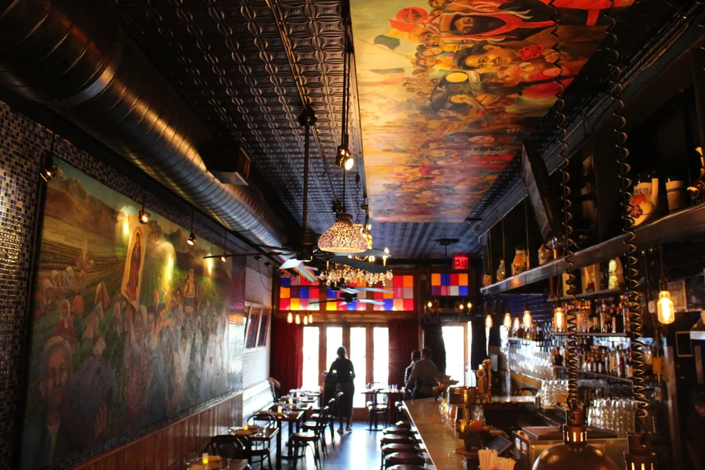 Restaurant interior with murals, patterned ceiling, exposed ducts, and bar.