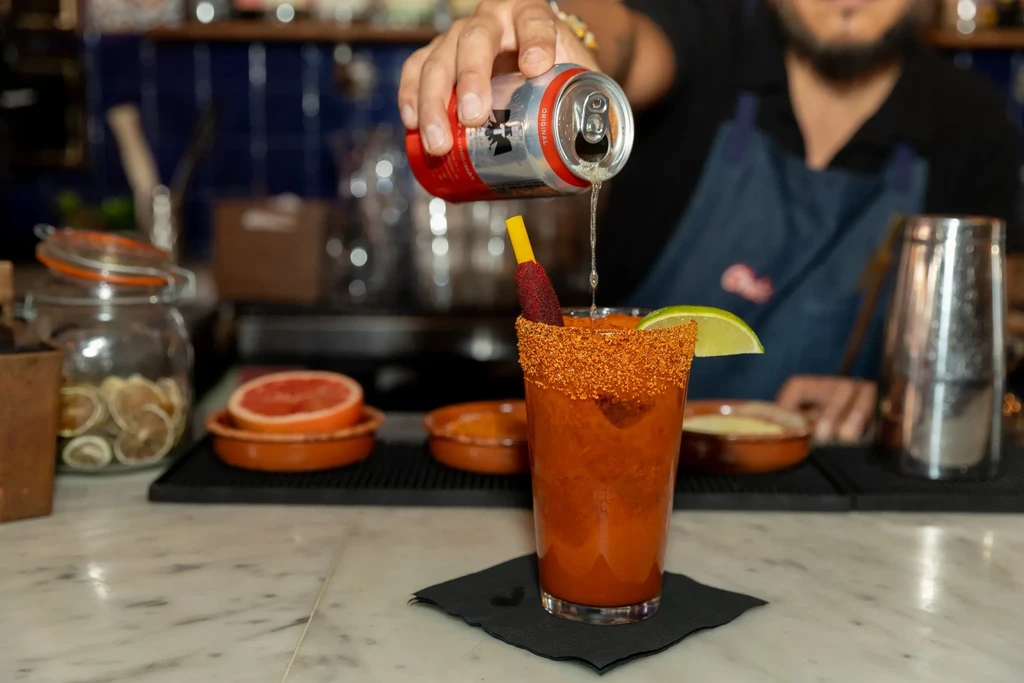 A bartender pours a drink from a can into a chili-rimmed glass.