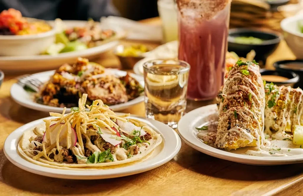 A table laden with Mexican food including tacos, elote, and drinks.