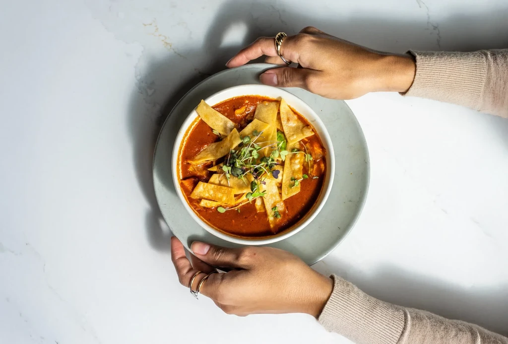 Hands serve a bowl of red soup with tortilla strips and sprouts.