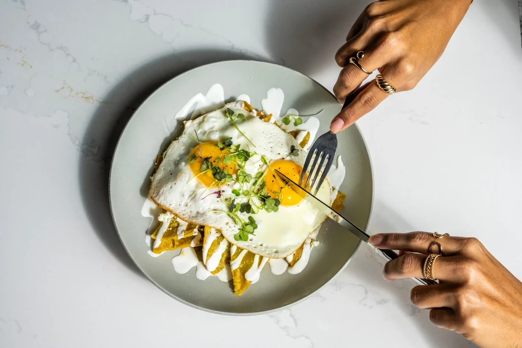 Hands cutting into sunny-side-up eggs and tortilla chips.