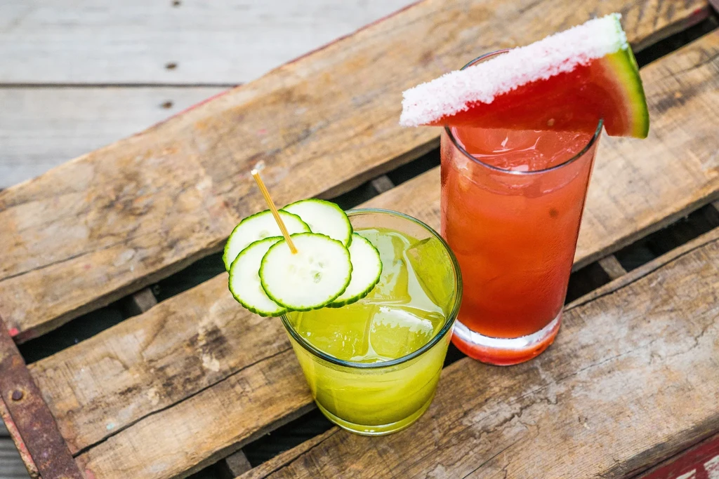 Two cocktails, one with cucumber, one with watermelon, on a wooden surface.