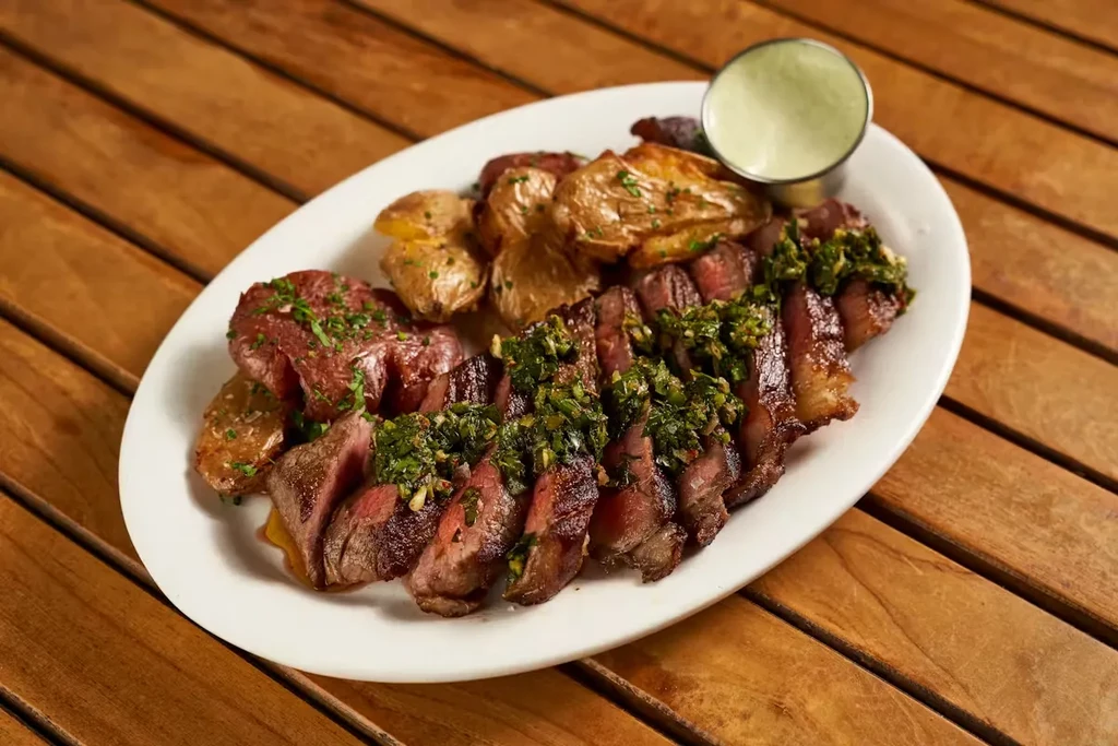 Grilled steak with chimichurri sauce, roasted potatoes, and a side of dressing.