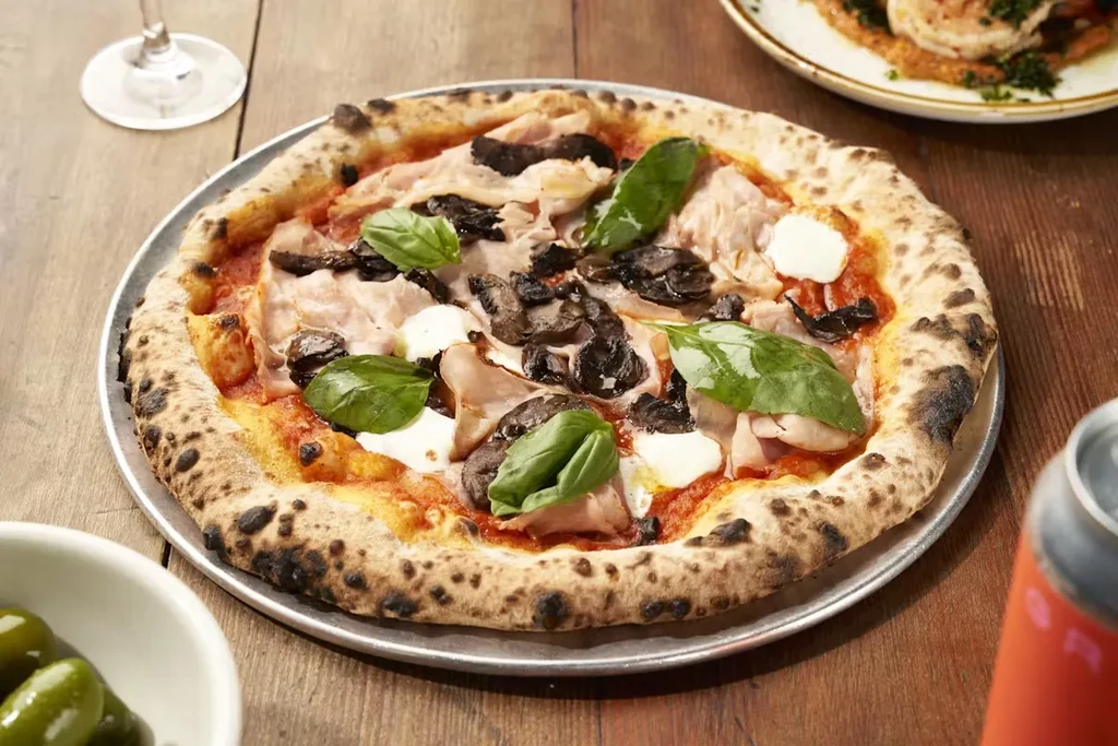 Neapolitan pizza with ham, mushrooms, mozzarella, and basil on a wooden table.