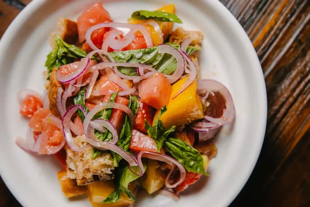 Panzanella salad with watermelon, tomatoes, red onion, and basil in a white bowl.