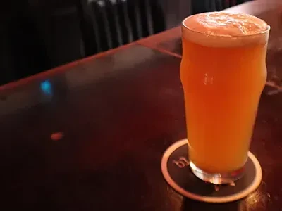 A glass of pale orange beer with foam on a dark bar.