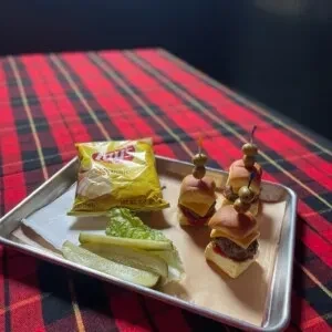 Sliders, chips, and pickles on a tray over a red plaid tablecloth.