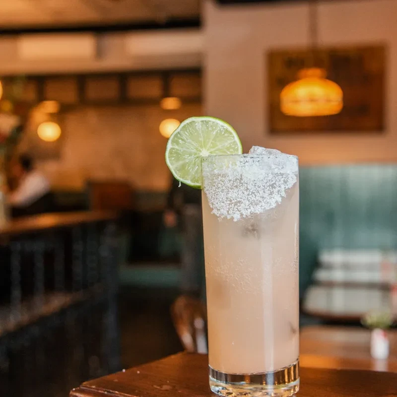 A tall, light-colored cocktail with salt rim and lime wedge.