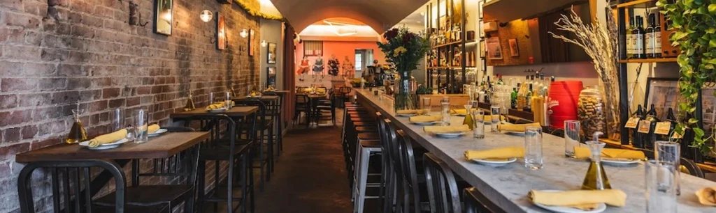 An empty restaurant interior with a long bar and tables along a brick wall.