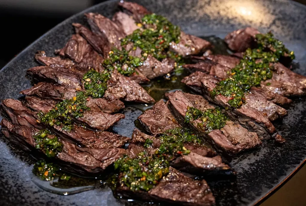 Sliced beef steak with a green herb sauce on a dark plate.