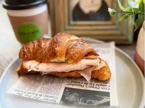 A croissant sandwich with ham on a newspaper with a coffee cup.