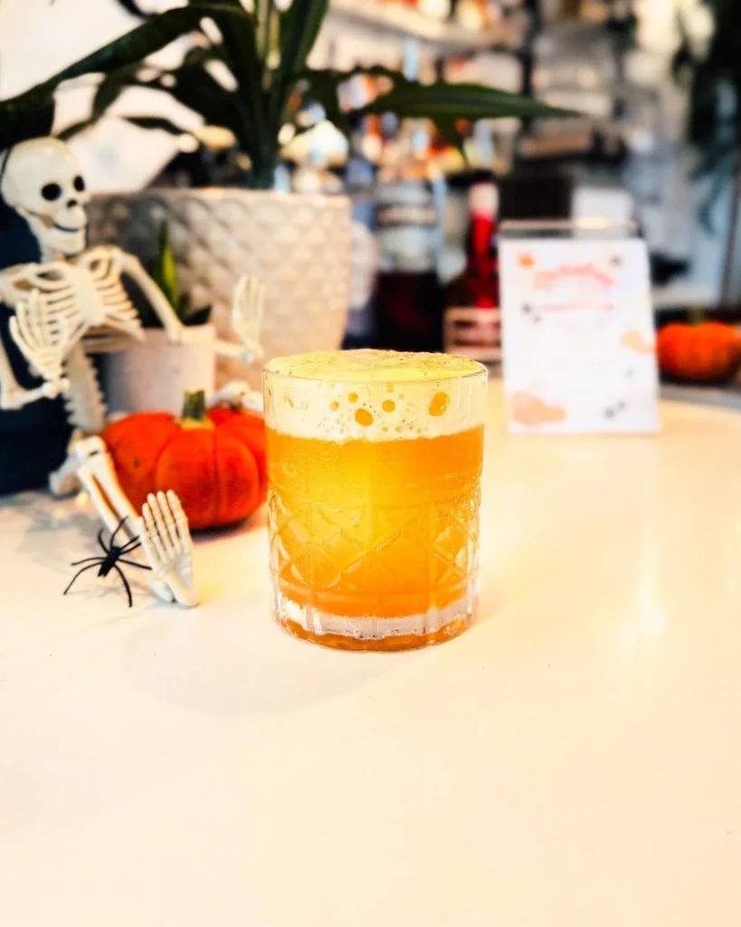 A foamy orange drink in a faceted glass with Halloween decorations.