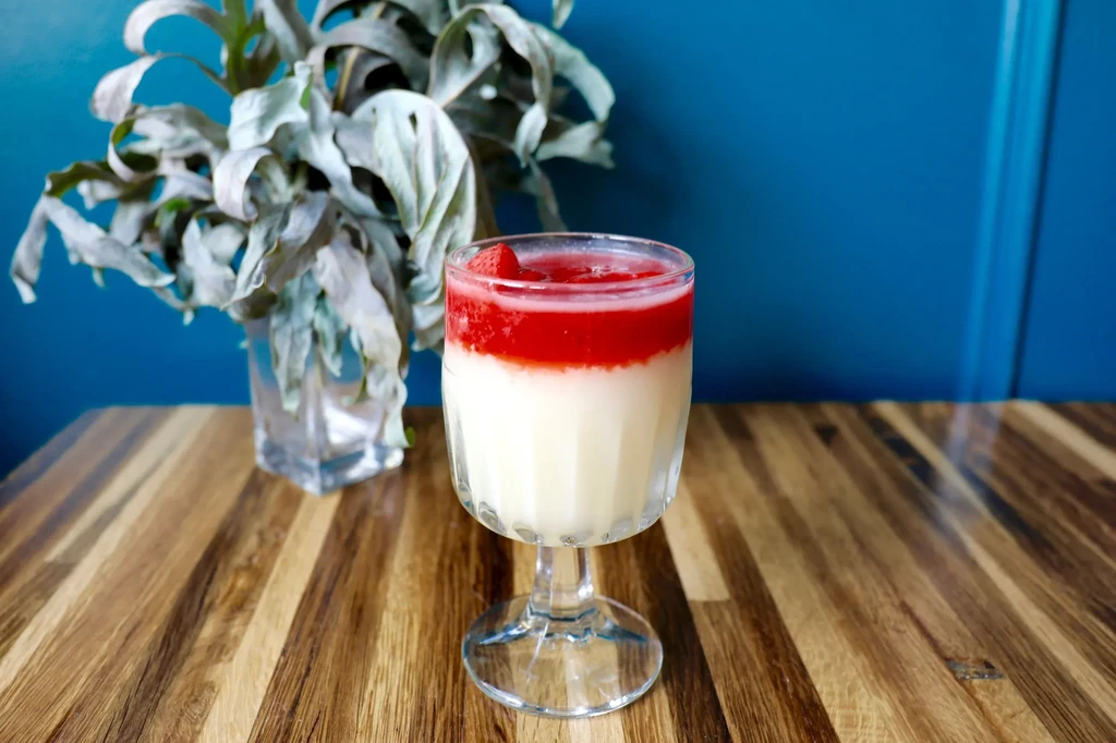 A glass of panna cotta with strawberry coulis on a wooden table.