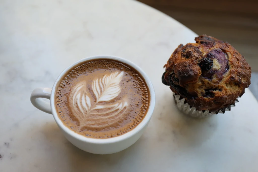 A cup of latte with leaf art and a blueberry muffin on marble.