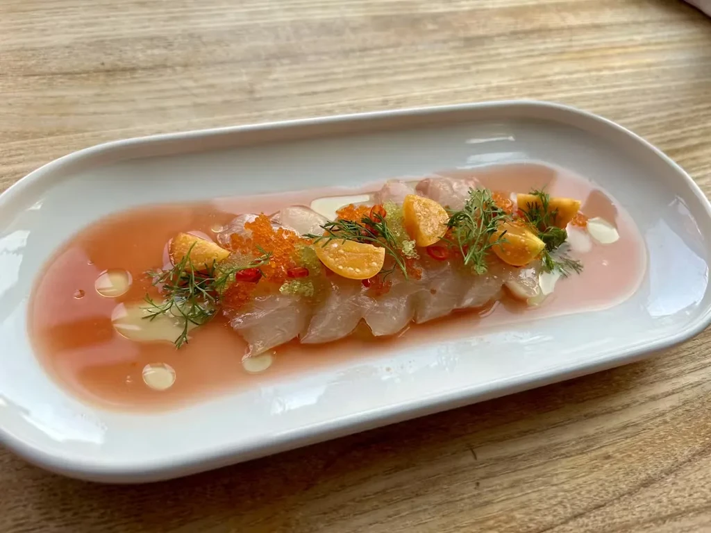 White plate of sliced fish with orange sauce, caviar, and herbs.