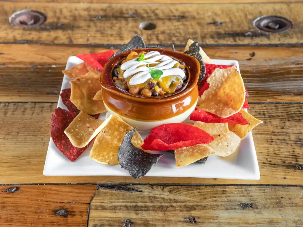 A brown bowl of chili with sour cream and a side of multi-colored tortilla chips.