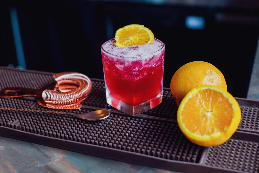 A red cocktail with an orange slice and bar tools on a mat.