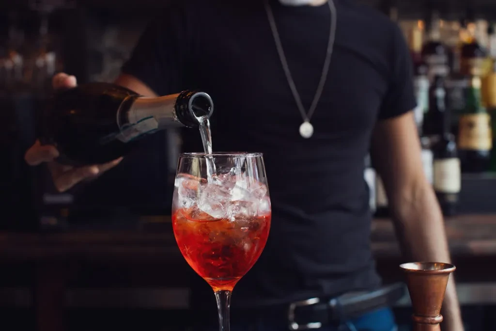 Bartender pouring clear liquid from a bottle into a red cocktail.