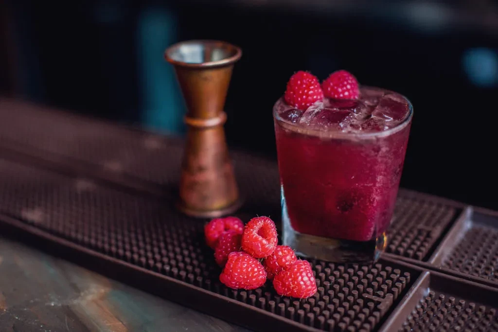 A raspberry cocktail with ice and a bronze jigger on a bar mat.
