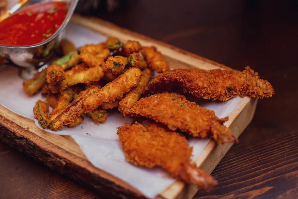A wooden tray with crispy shrimp, fried zucchini, and a red dipping sauce.