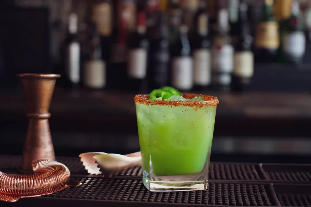 A green cocktail with a chili rim and jalapeño garnish at a bar.