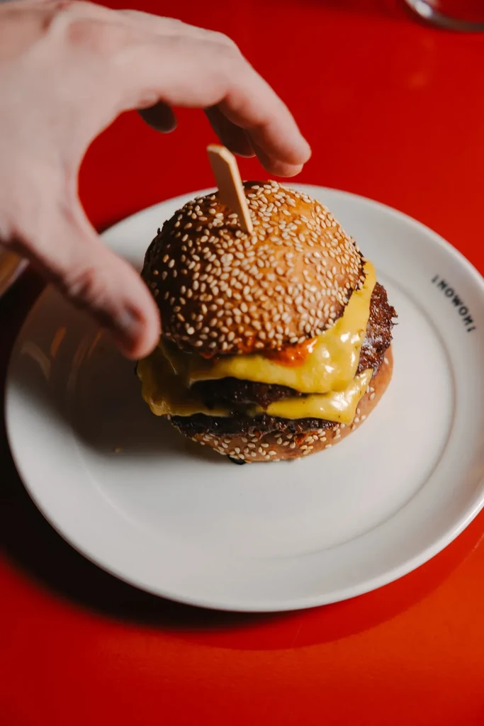 A hand is about to grasp a double cheeseburger on a white plate.