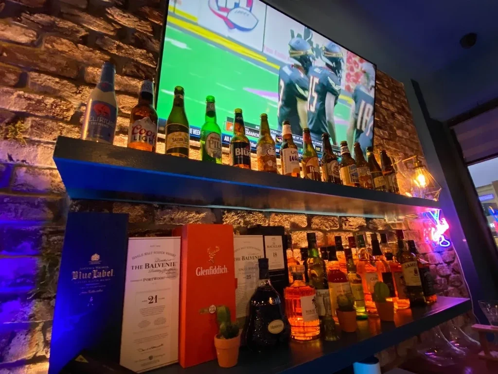 A bar with alcohol bottles, a TV showing football, and brick wall.