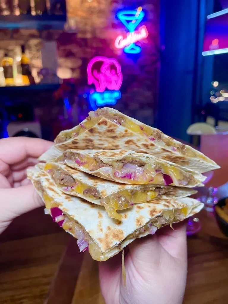 Hands hold quesadilla slices with melted cheese and red onion under neon lights.