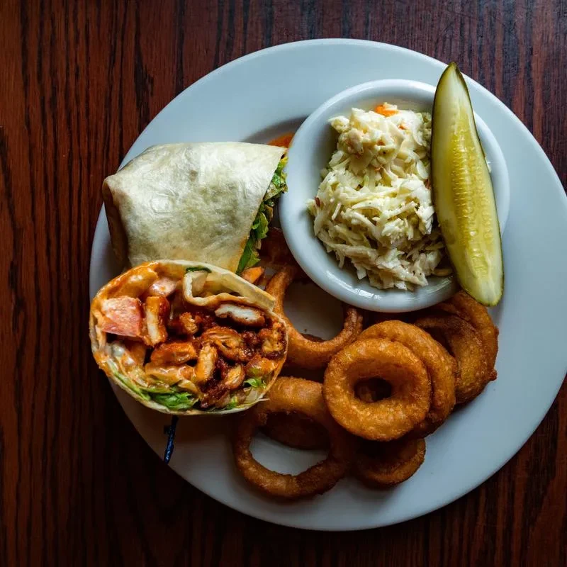 A chicken wrap, onion rings, coleslaw, and a pickle on a white plate.
