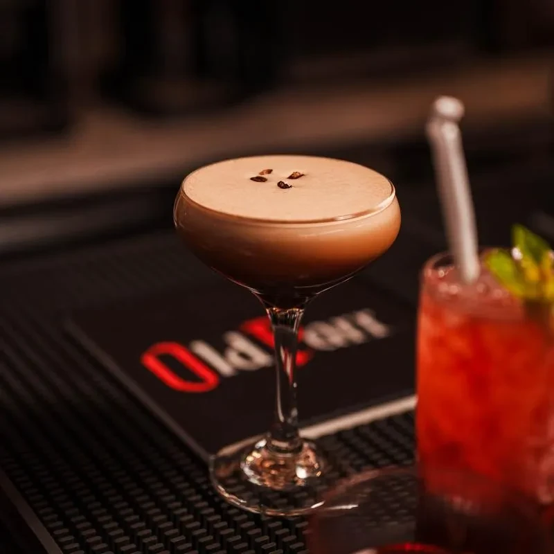 Espresso martini with foam and coffee beans on a dark bar mat.