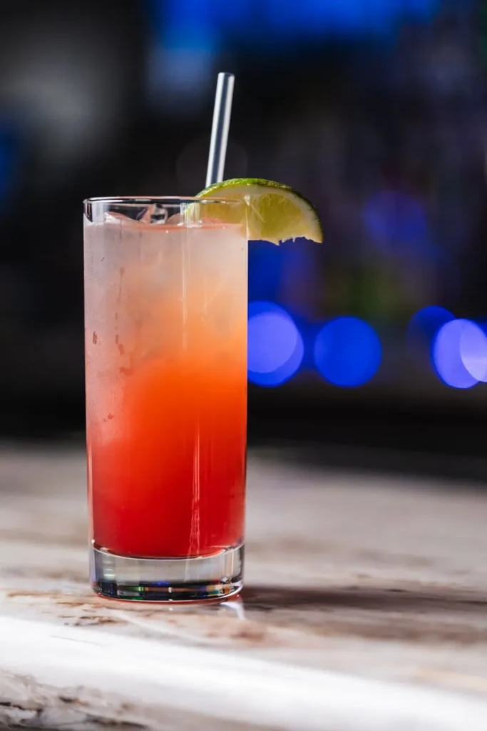 Tall glass with layered red and orange drink, lime, straw, ice.