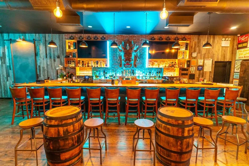 An empty rustic bar with stools, wooden barrels, and blue neon lighting.