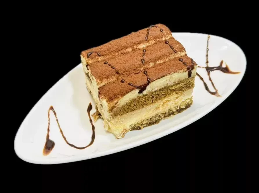 A slice of layered tiramisu cake with cocoa powder and chocolate drizzle.