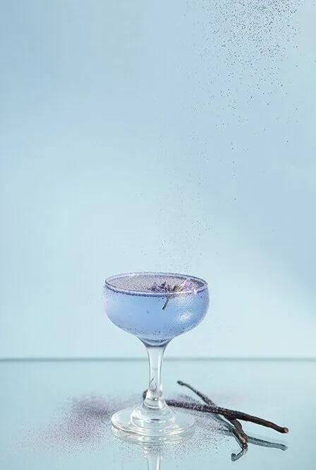A coupe glass with purple liquid, flower garnish, and falling glitter.