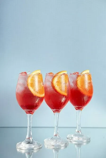 Three glasses of red aperitif cocktails with orange slices against a blue background.