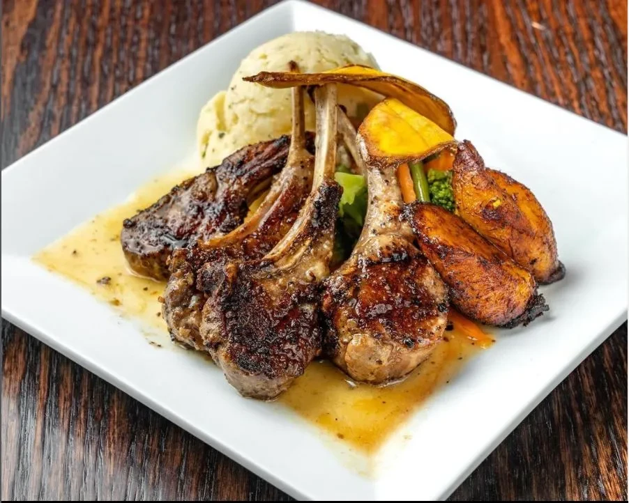 Grilled lamb chops with mashed potatoes, vegetables, and plantains on a white plate.