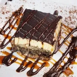 A slice of tiramisu with chocolate and caramel drizzle on a white plate.