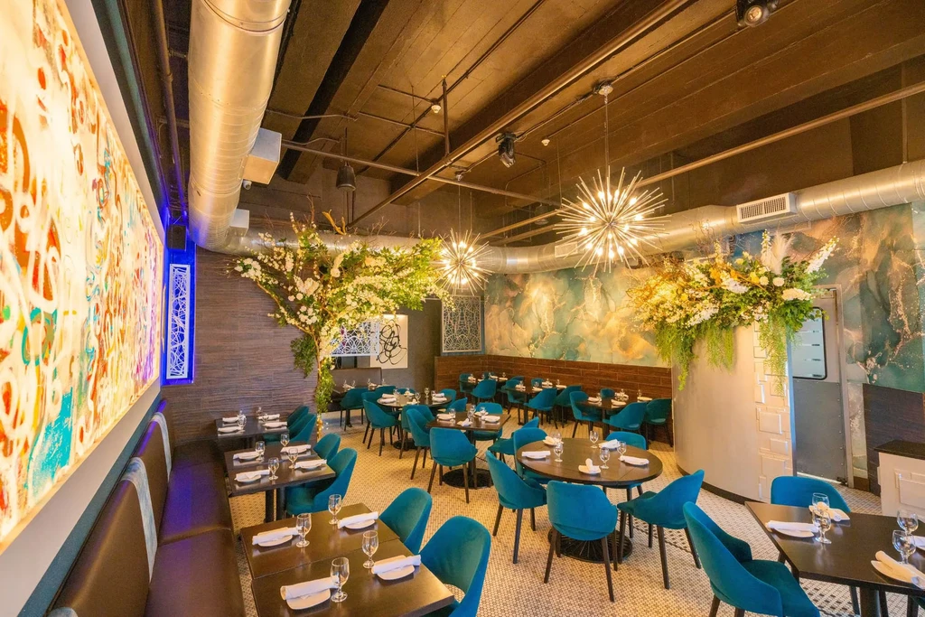 A modern restaurant interior with teal chairs, flower decor, and unique lighting.