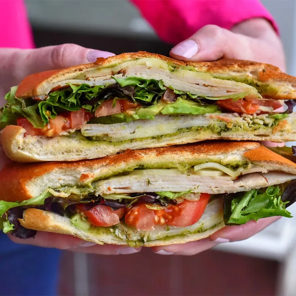 Hands hold a sliced turkey panini with pesto, lettuce, tomato, and avocado.