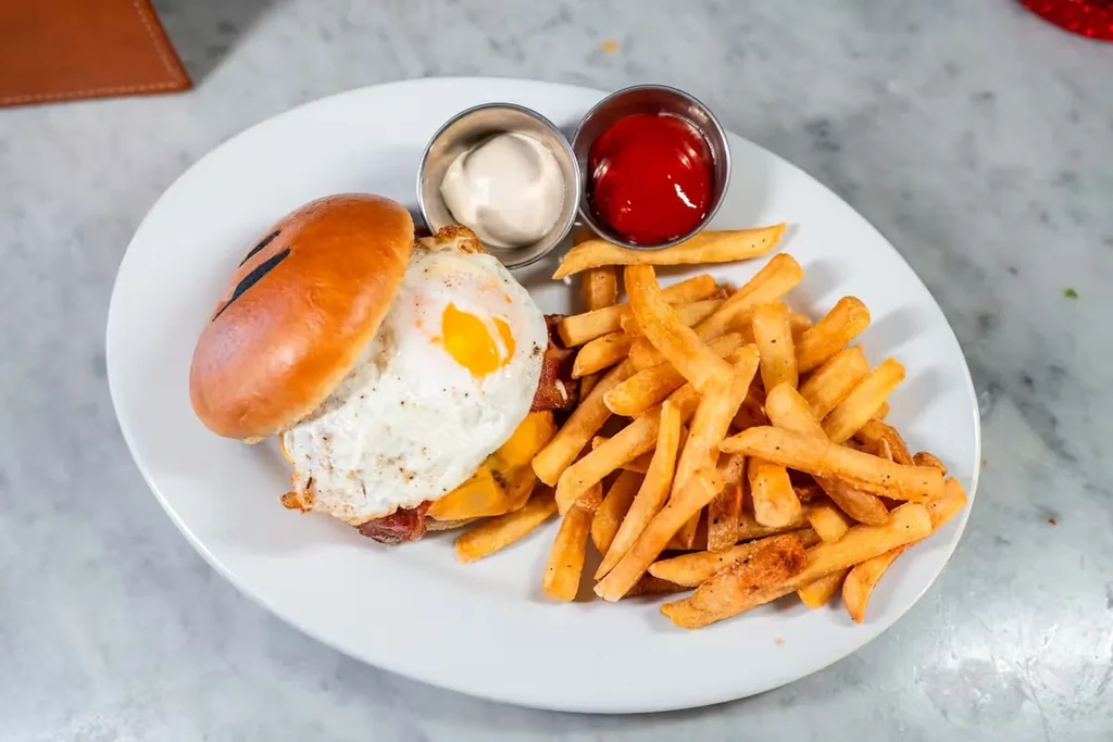 A fried egg burger with fries, ketchup, and mayonnaise on a white plate.