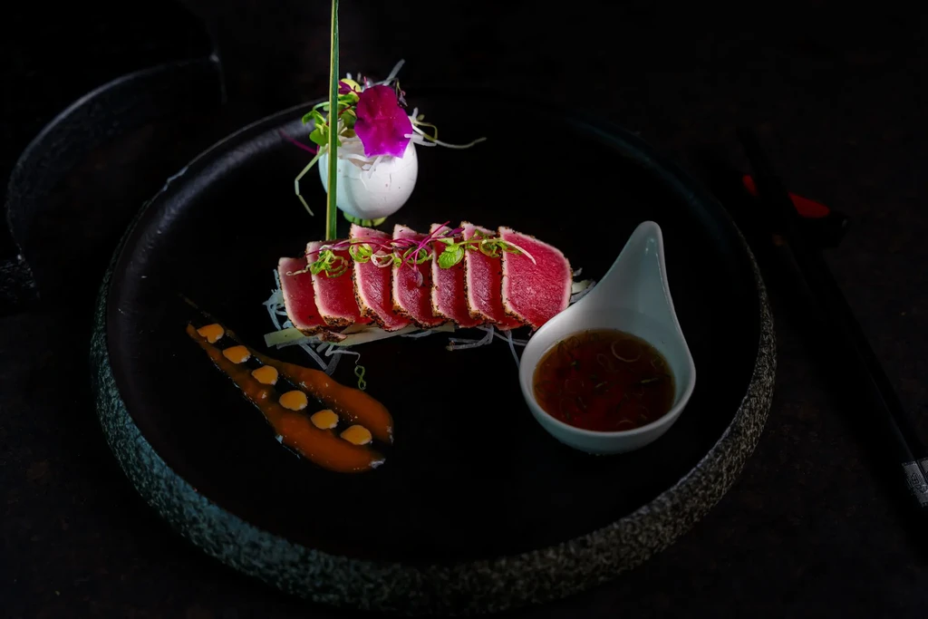 Seared tuna slices with garnish and dipping sauce on a black plate.