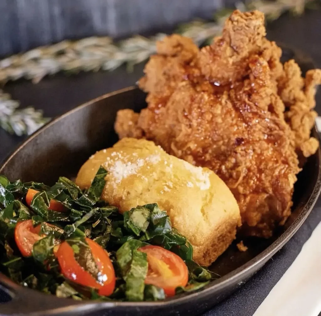 Fried chicken, cornbread, and greens with tomatoes in a black bowl.