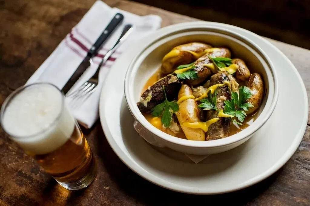 A bowl of sausages and potatoes with mustard and parsley, with a beer.