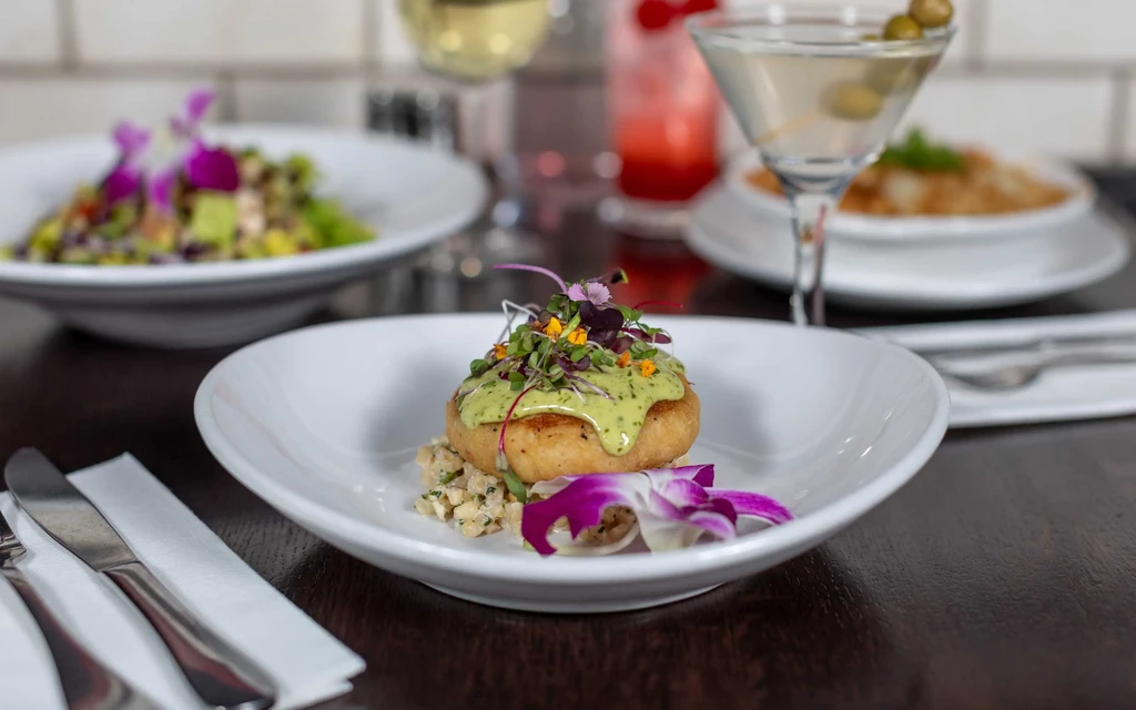 A golden-brown crab cake with green sauce, microgreens, and a purple orchid.
