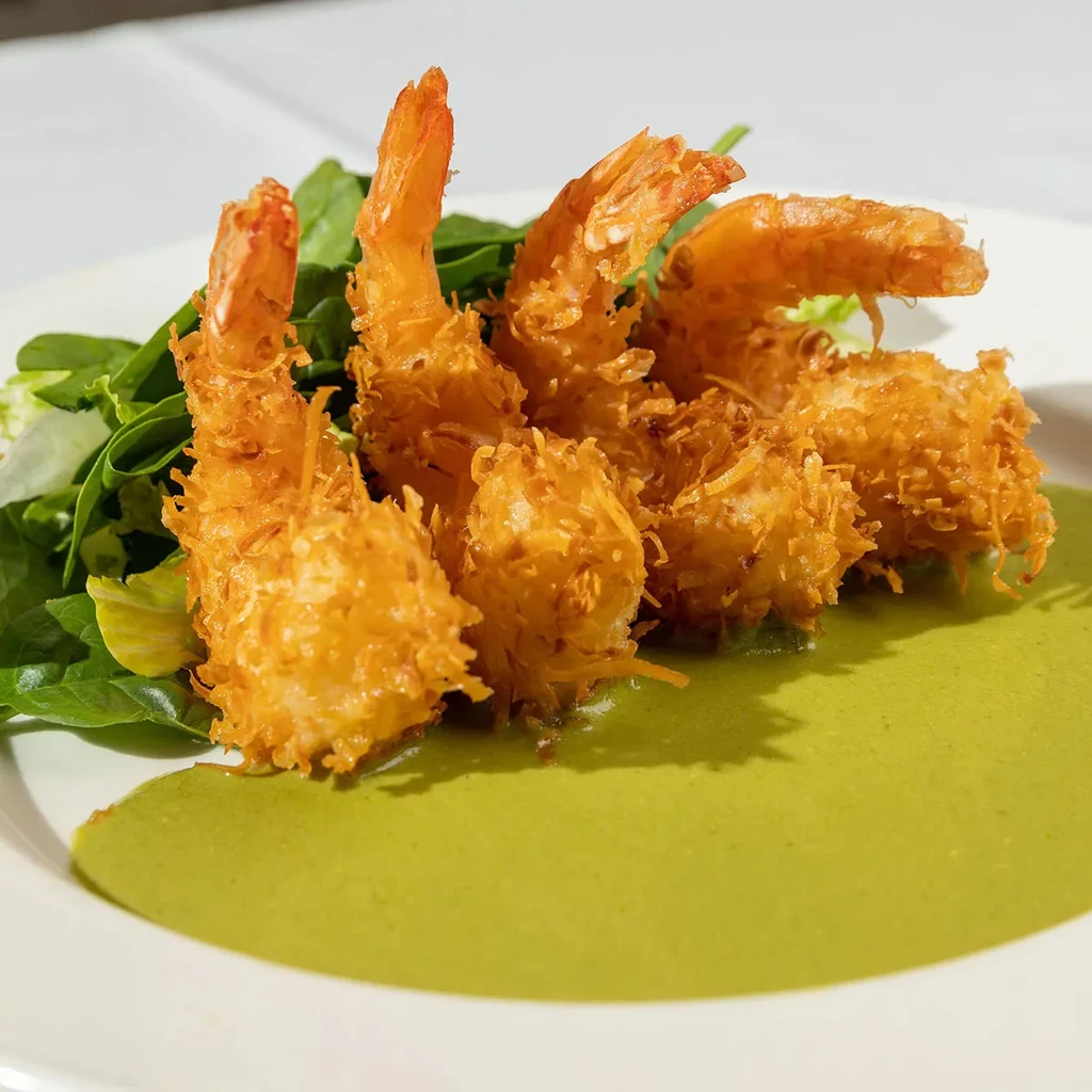 A plate of crispy coconut shrimp with green sauce and greens.
