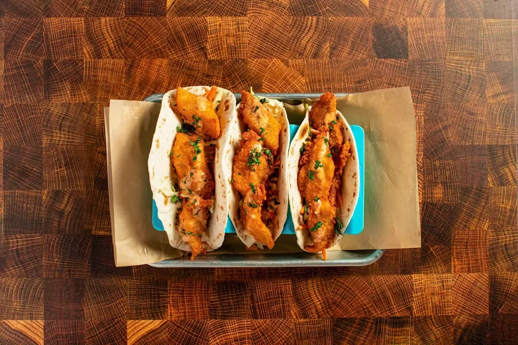 Three fried fish tacos with sauce and cilantro on a wood table.