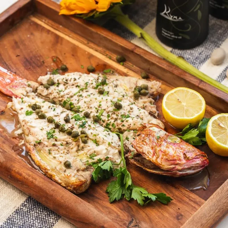 Two fish fillets with capers and parsley, lemon halves on a wooden board.
