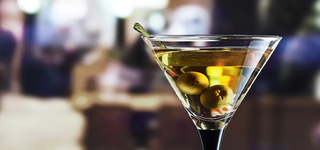 A martini glass with two olives and a stir stick against a blurred background.
