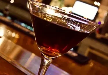 A dark-colored drink in a martini glass on a blurred bar background.