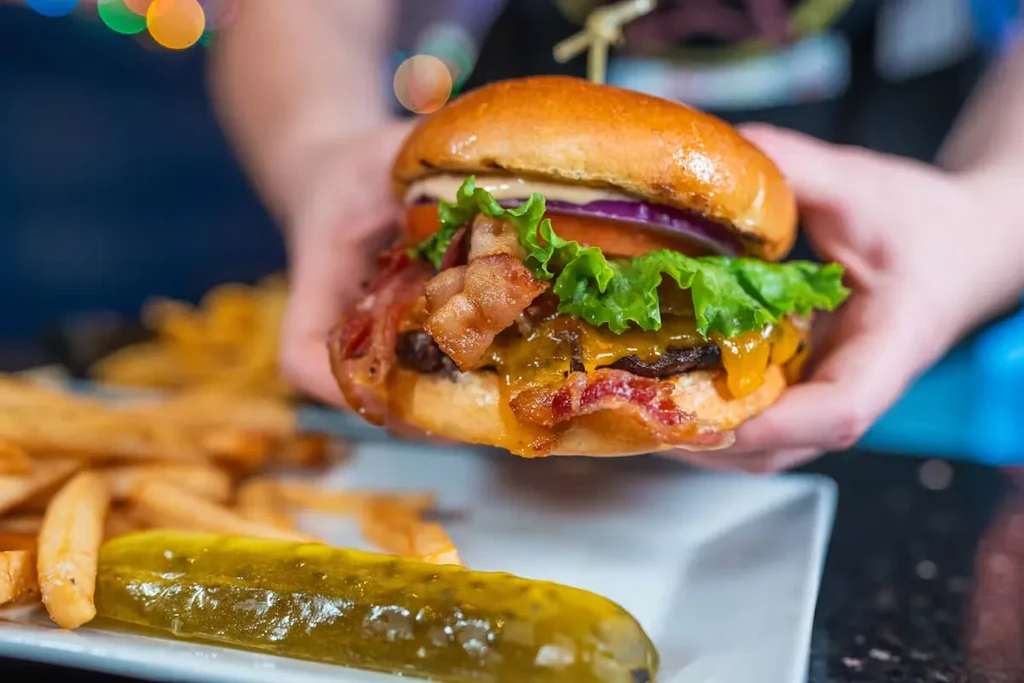 Hands holding a bacon cheeseburger with fries and a pickle.